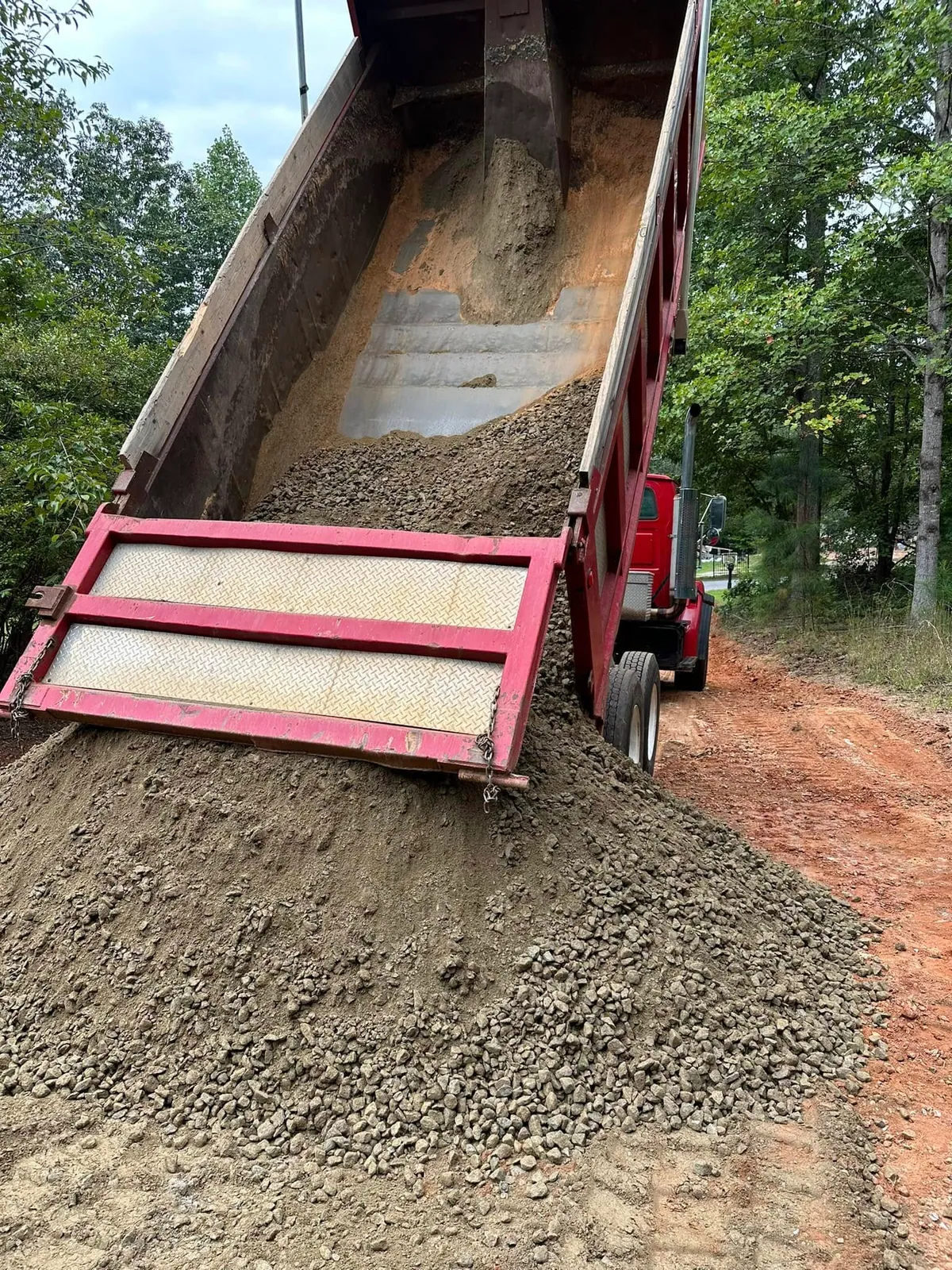 Red tandem dump truck with bed raised dumping crusher-run gravel onto wooded driveway